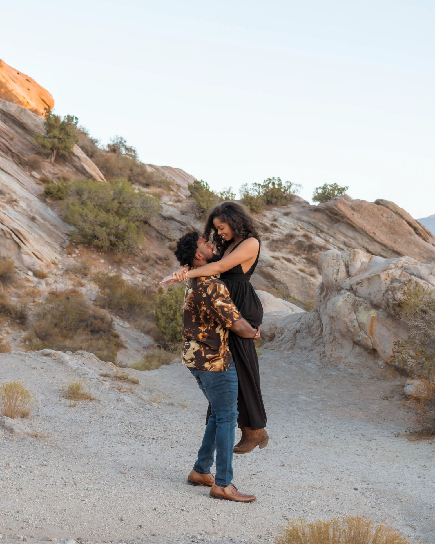 Couple embracing and dancing in a rocky desert landscape with sandy terrain and sparse desert vegetation.