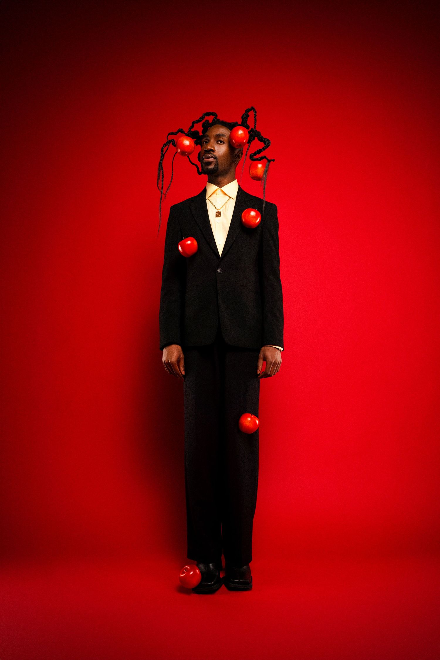 Artistic portrait against red backdrop featuring black suit and floating red apples intertwined with braided hair.
