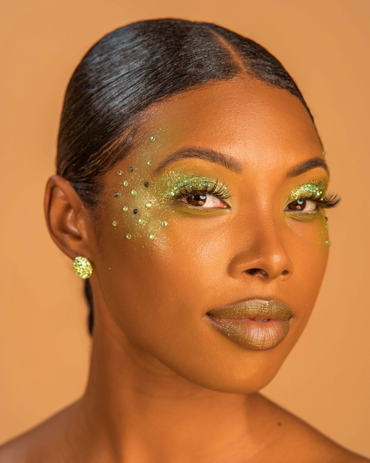 Beauty close-up featuring glittery gold eye makeup and sleek hairstyle against orange background.