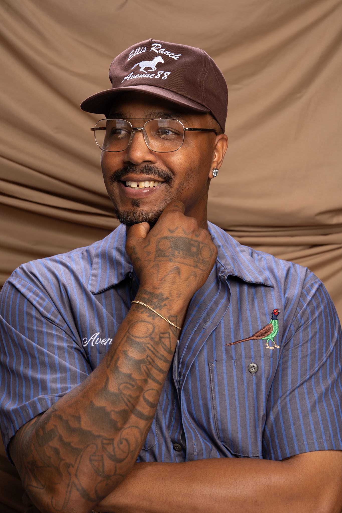 A person wearing a brown baseball cap and blue striped work uniform smiles while showing off their tattooed arm.