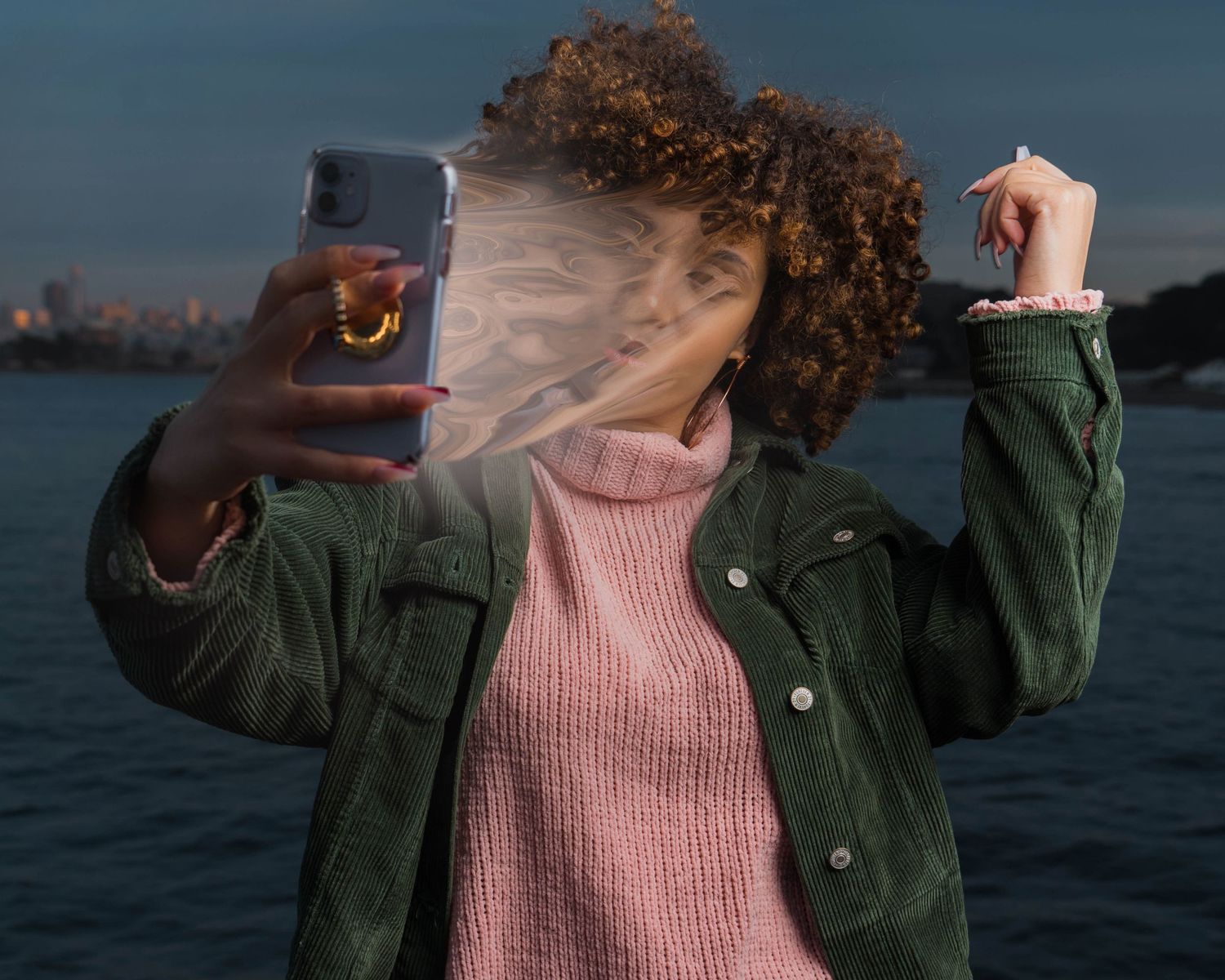 Person in green jacket and pink sweater posed against waterfront backdrop at dusk.