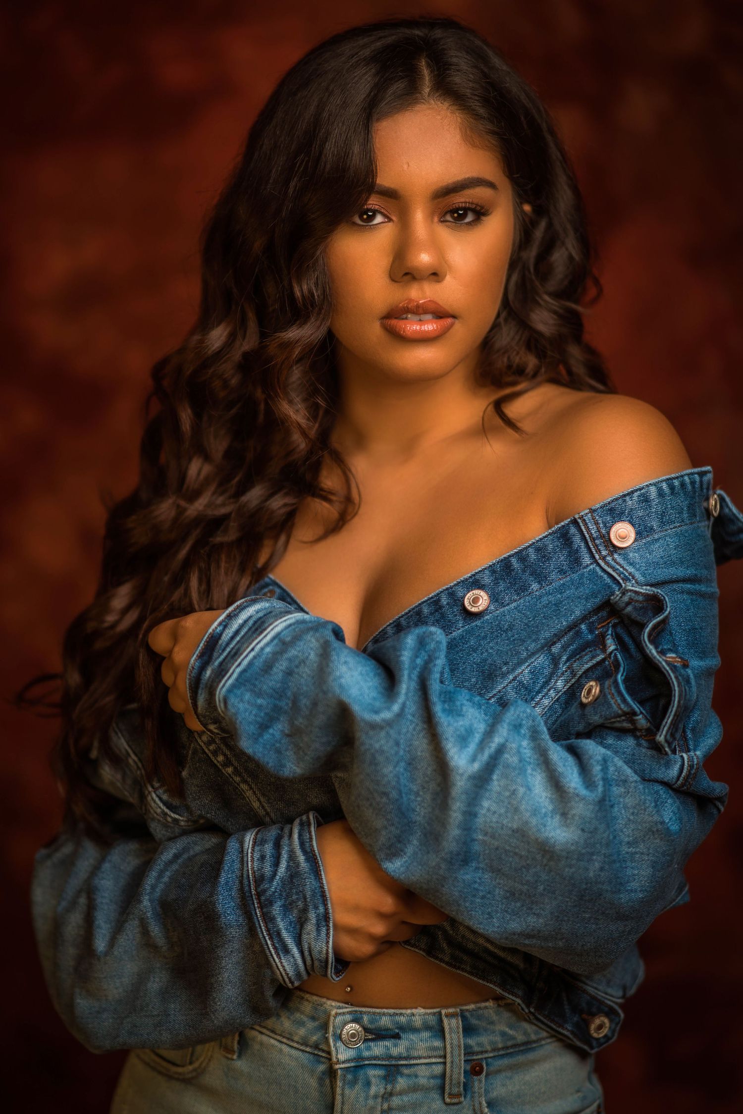 Young woman with long curly hair poses confidently in an off-shoulder denim jacket and jeans against a dark red background.