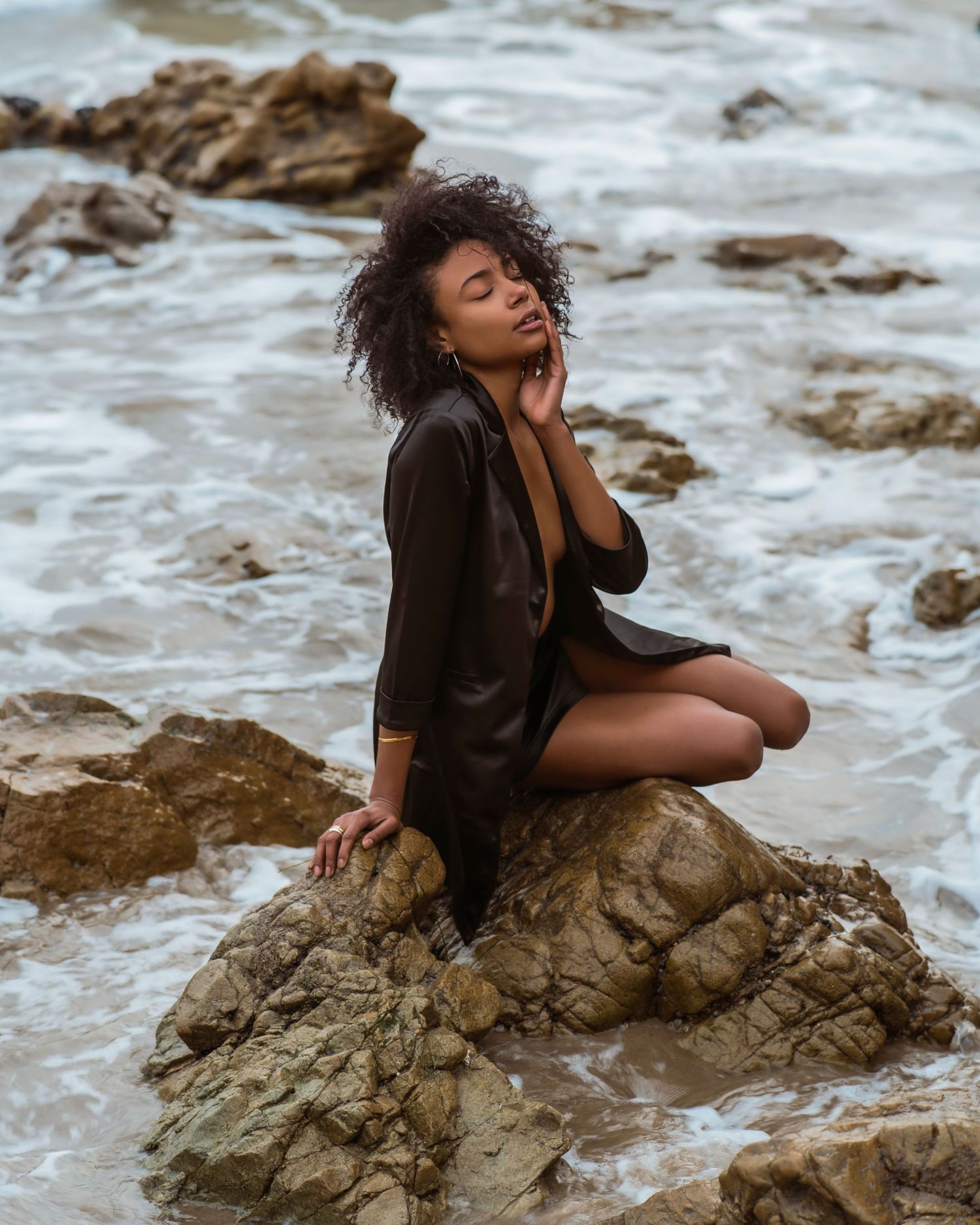 A woman in a black outfit sits gracefully on coastal rocks with eyes closed, ocean waves in the background.