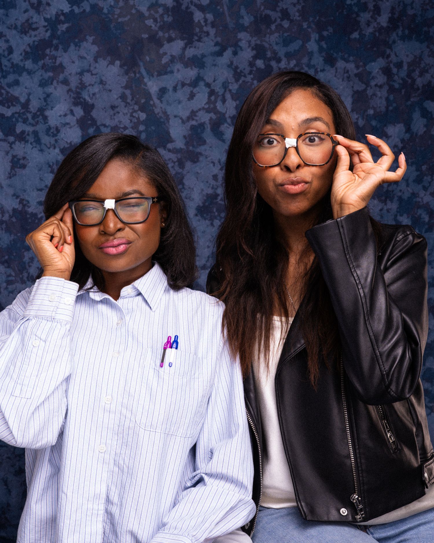 Two young women wearing oversized glasses pose playfully against a blue backdrop, one in a striped shirt and one in a black jacket.