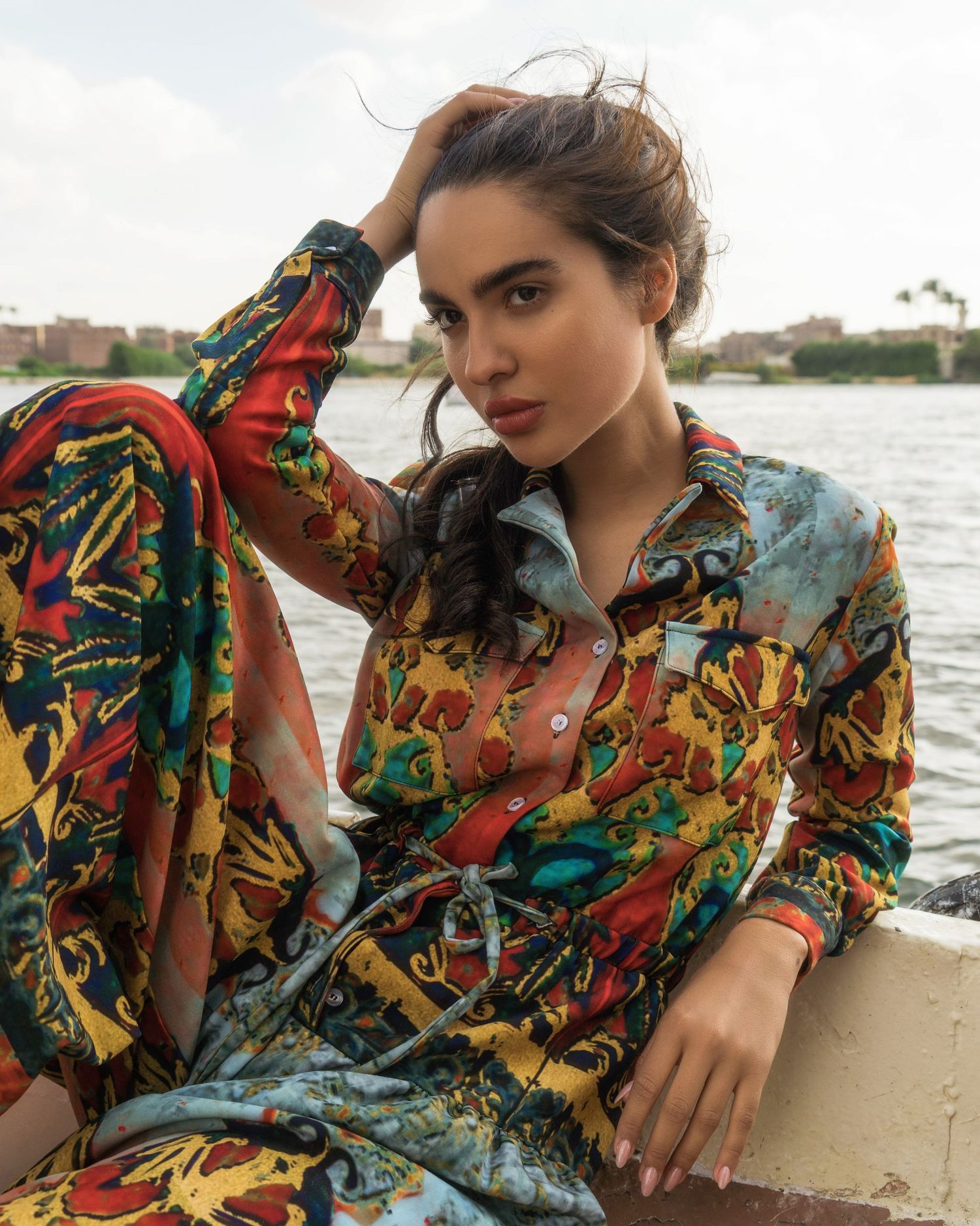 Model wearing colorful patterned dress lounging on boat deck with waterfront views.