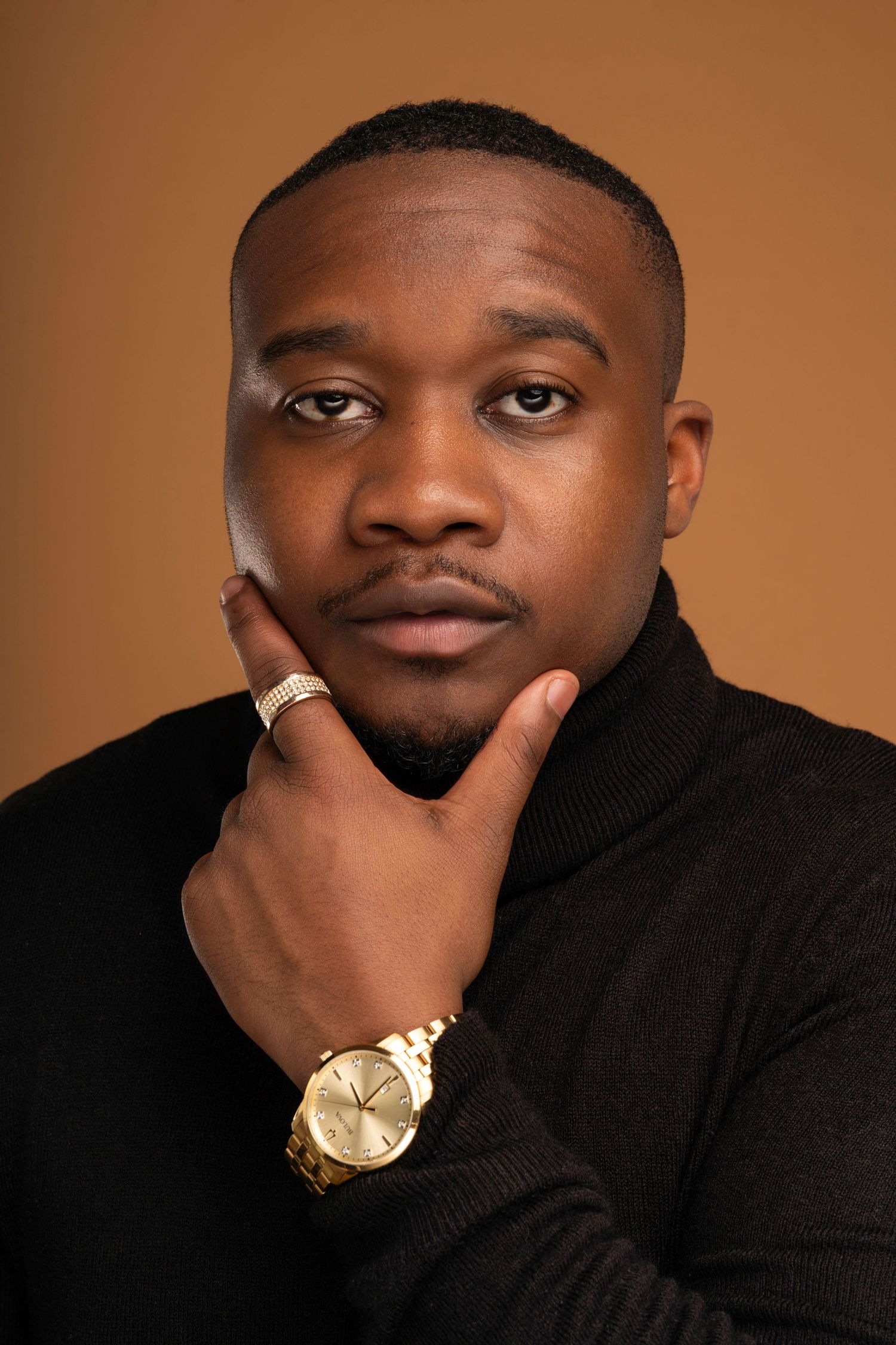 Professional headshot of a person in black turtleneck with gold watch against brown backdrop.
