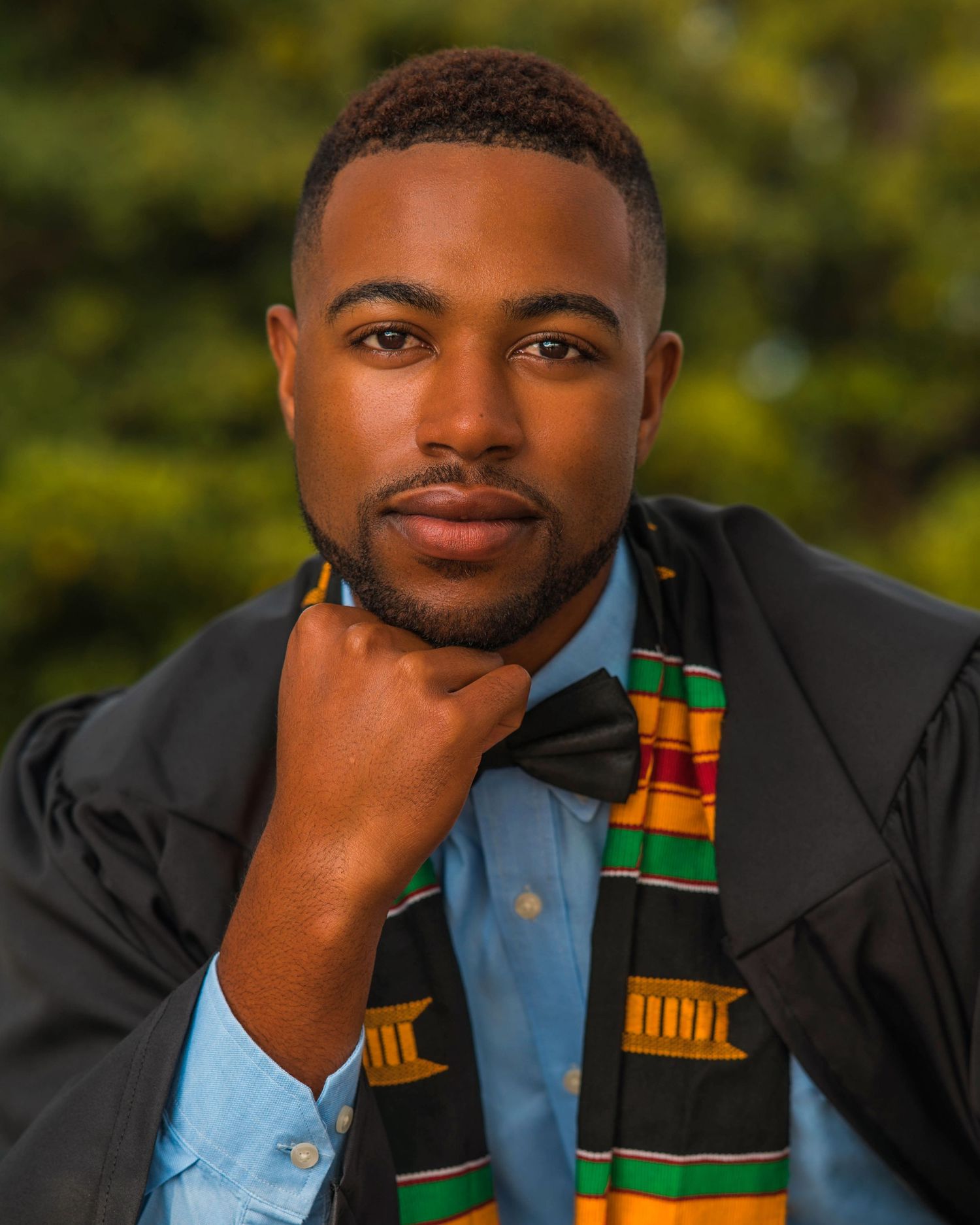 Young man in graduation gown and kente cloth stole poses confidently for senior portrait outdoors.