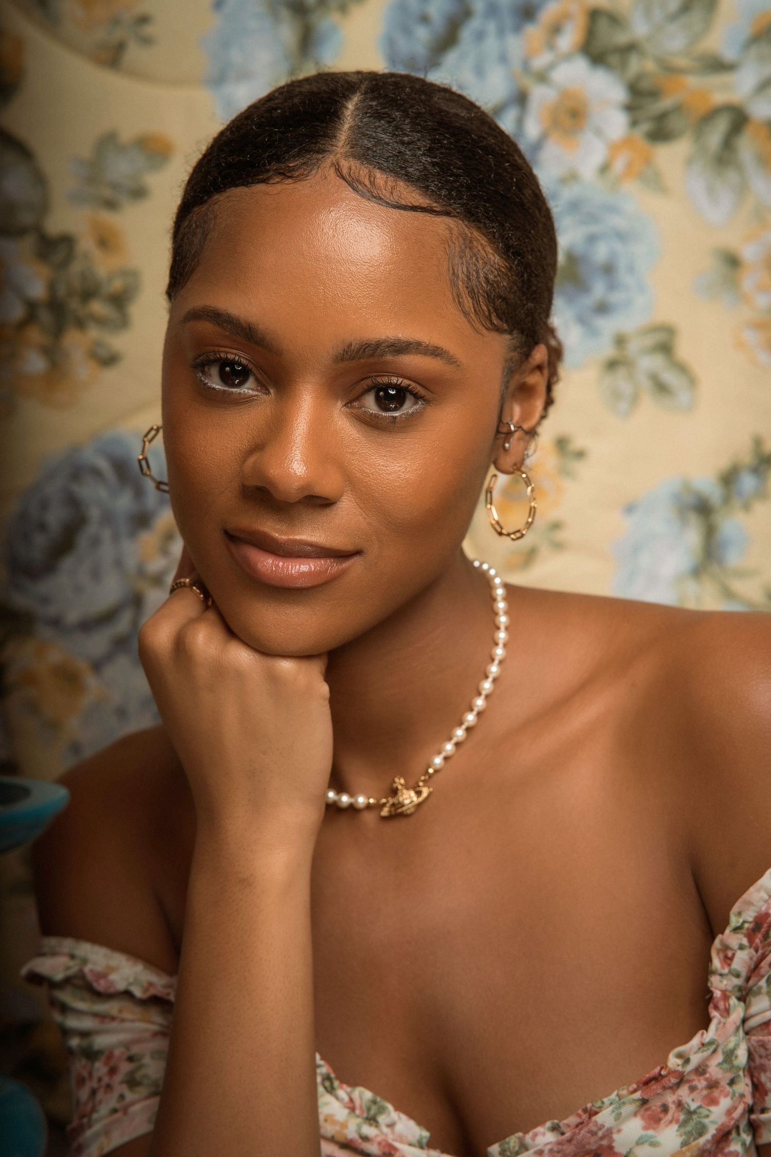 Portrait against floral wallpaper showcasing pearl necklace and delicate off-shoulder dress.