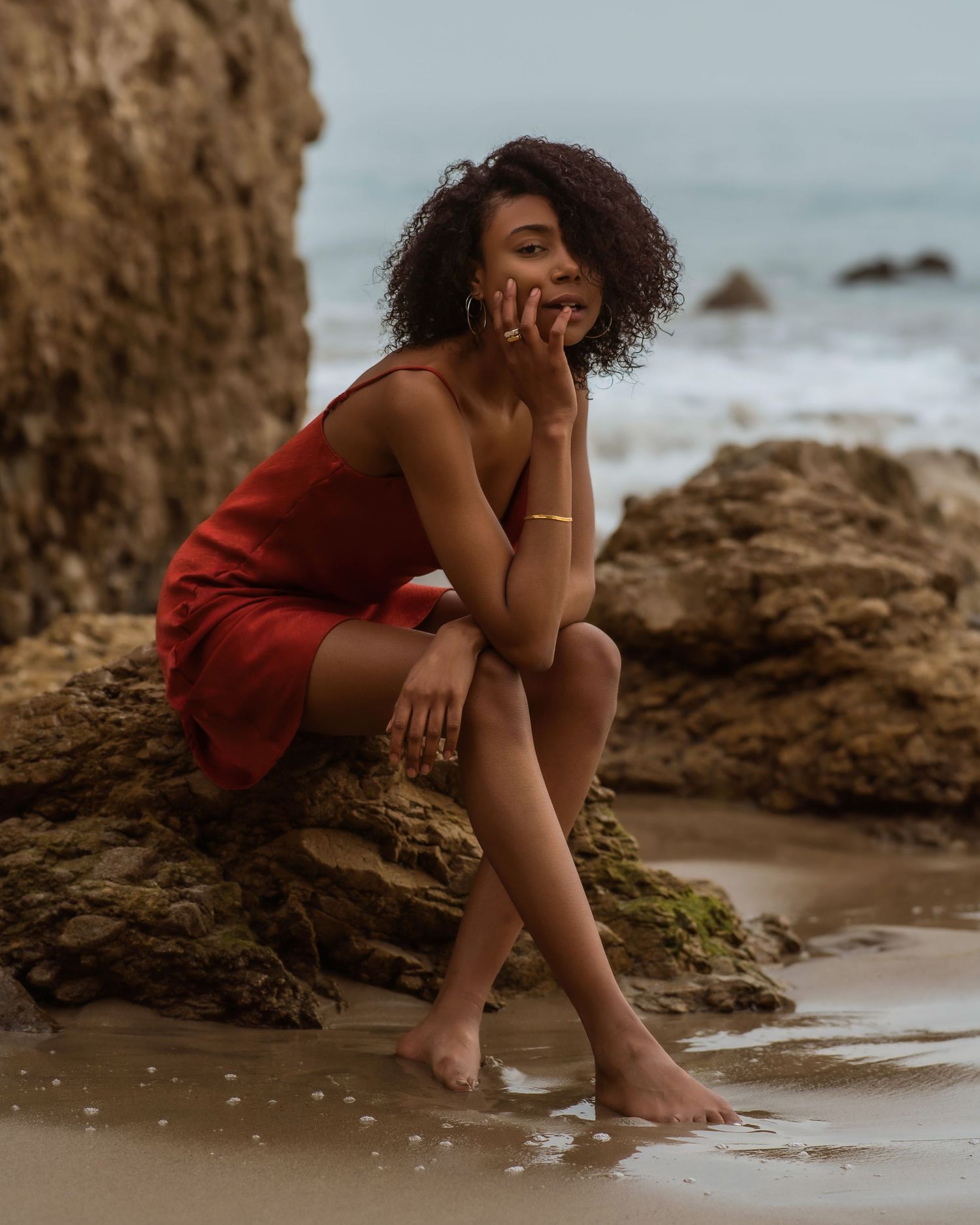 A young woman in a red dress sits barefoot on rocky beach, gazing thoughtfully with curly hair and gold jewelry.