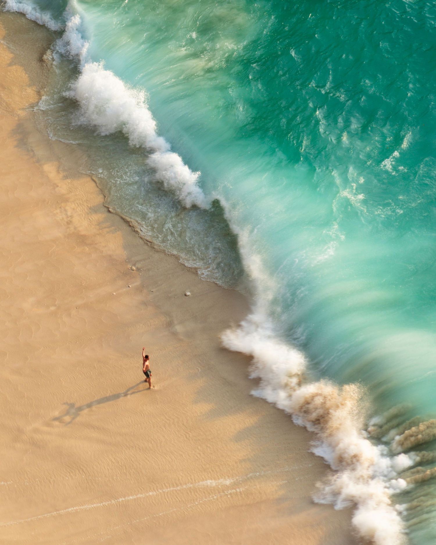 Bird's eye view of a lone figure walking along a beach with dramatic turquoise waves.