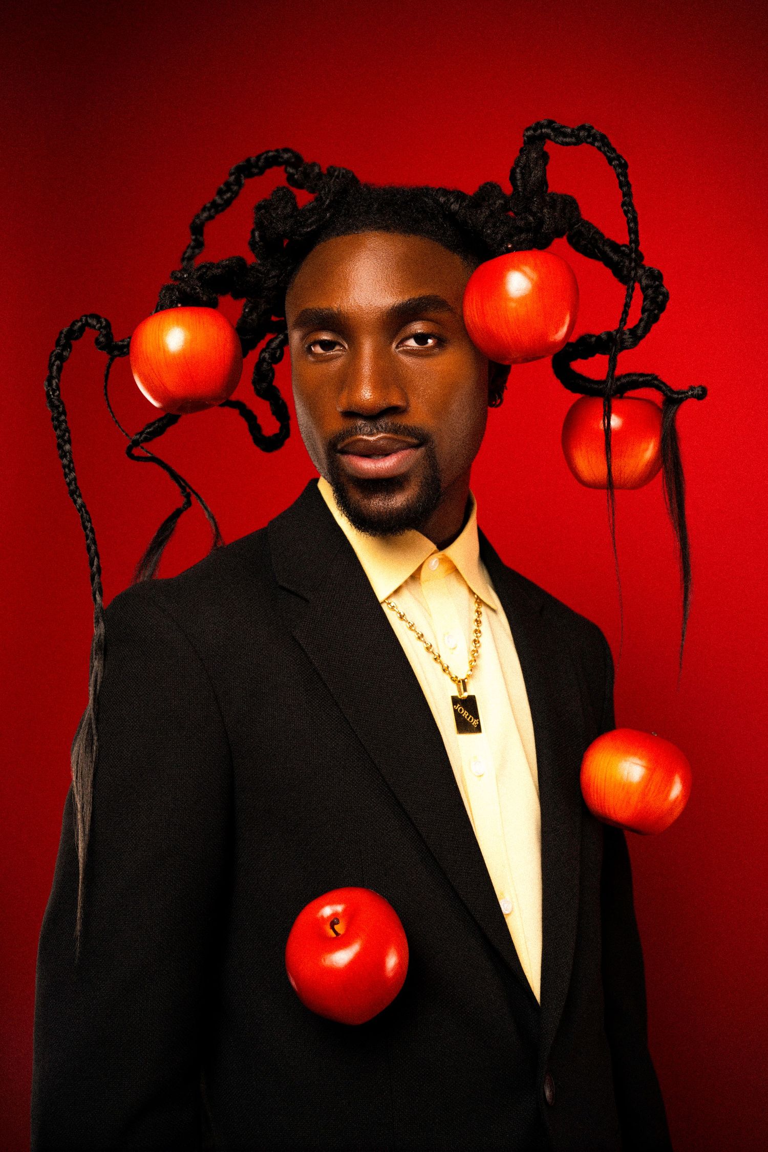 Elegant studio portrait with red apples suspended in braided hair wearing black suit and gold necklace.