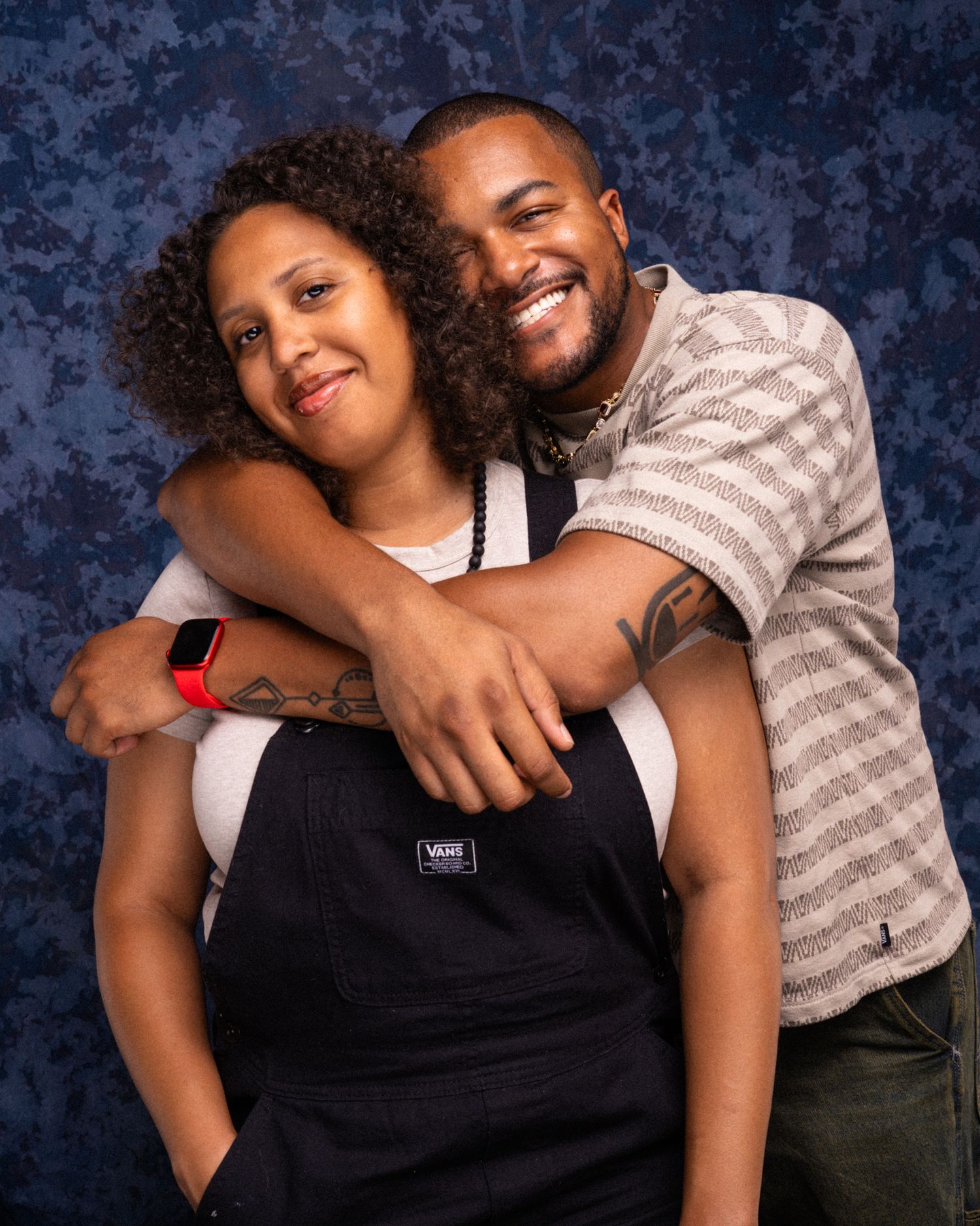 A smiling couple poses together, the man embracing the woman from behind, both wearing casual clothing against a blue backdrop.