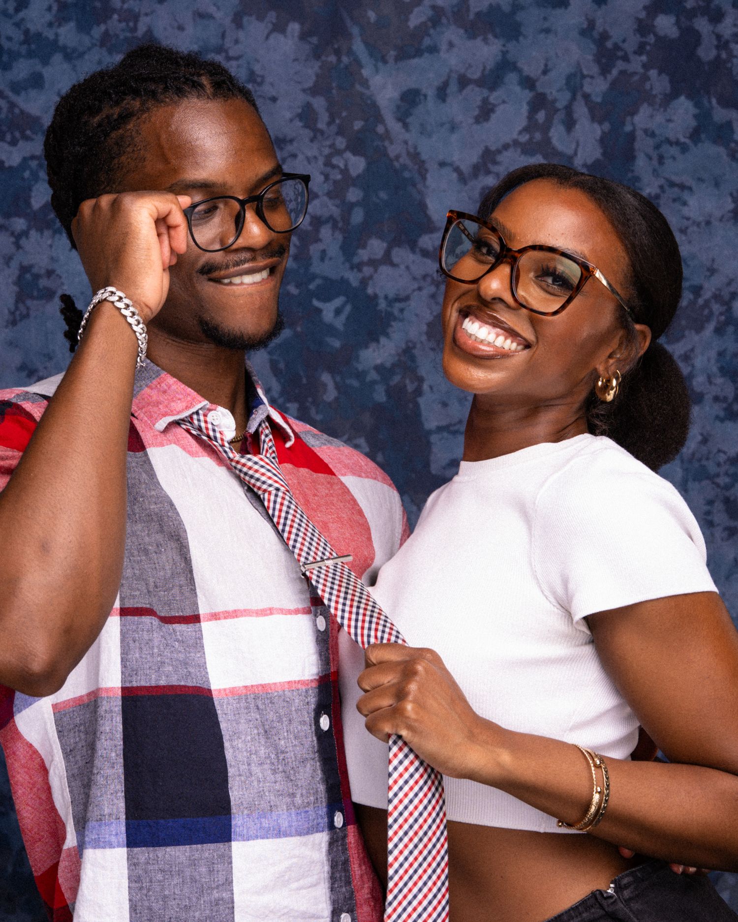 Two young adults wearing glasses and plaid ties pose playfully against a blue backdrop in matching outfits.