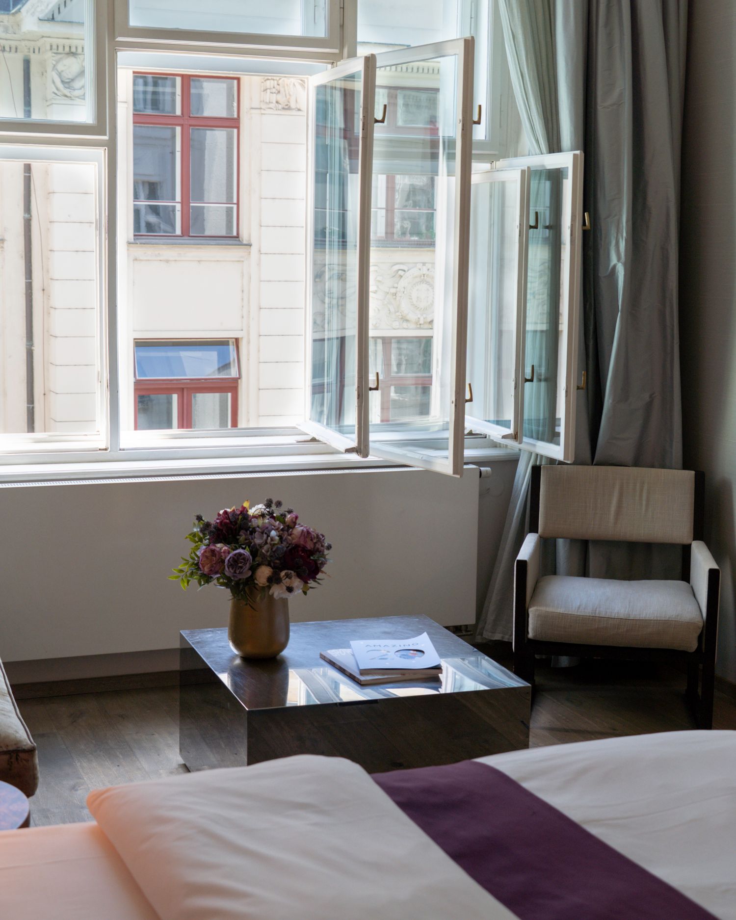 Elegant hotel room with open windows, fresh flowers, mirrored coffee table, and a cozy armchair overlooking a European street.