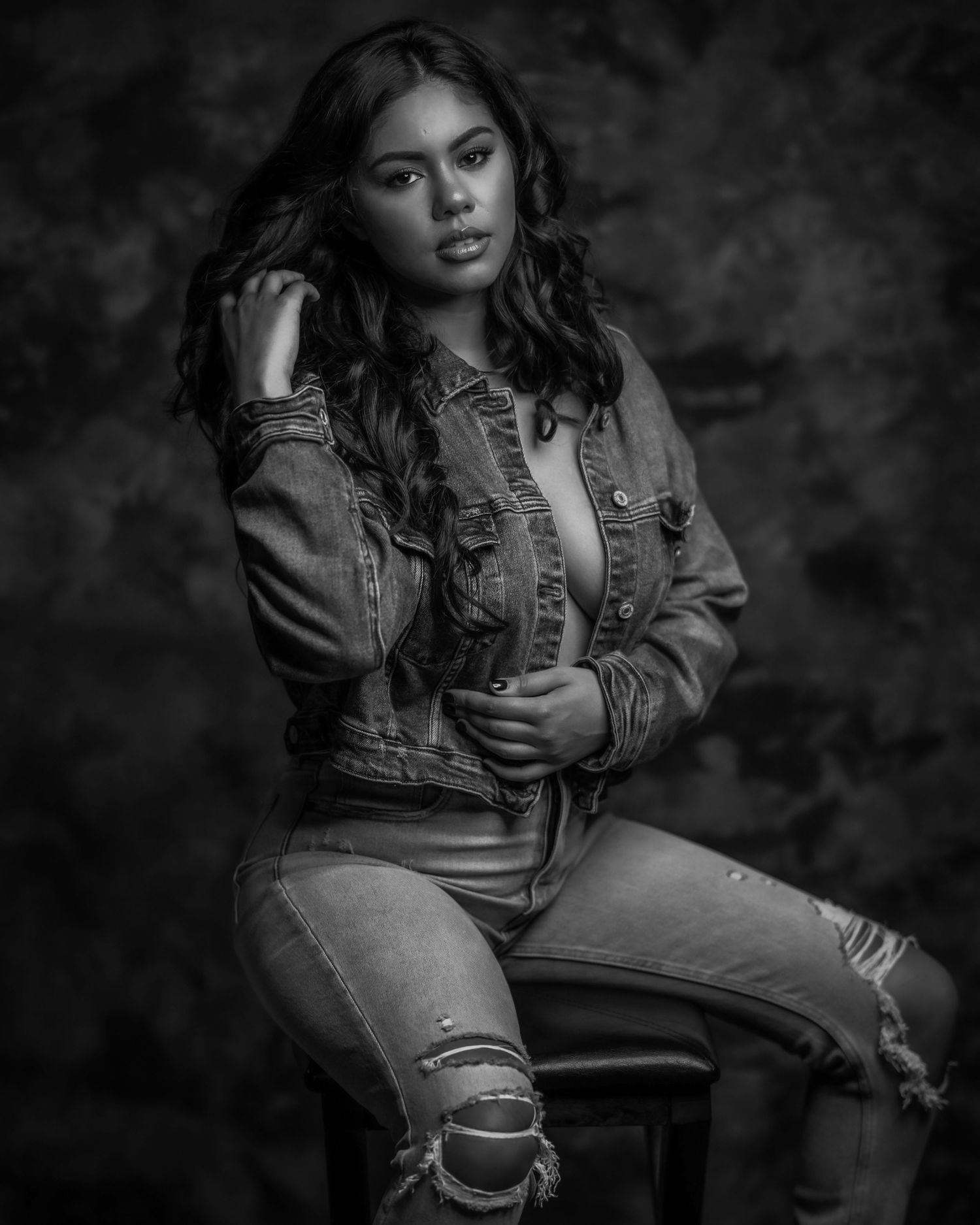 Black and white portrait of a young woman in a denim jacket and ripped jeans, posing confidently on a stool.