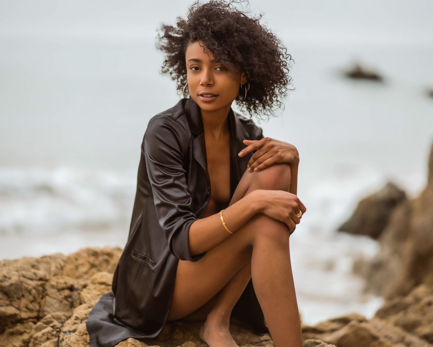 A young woman with curly hair sits on rocks at a beach, wearing a dark shirt, gazing thoughtfully at the camera.