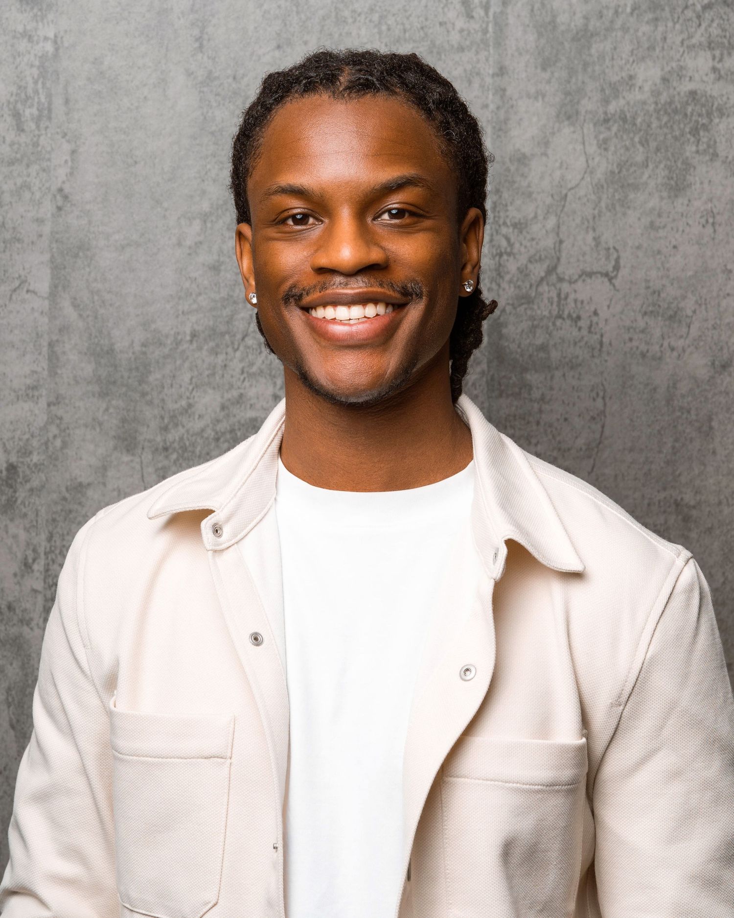 Professional headshot portrait of someone wearing a white jacket and t-shirt against a textured gray background.