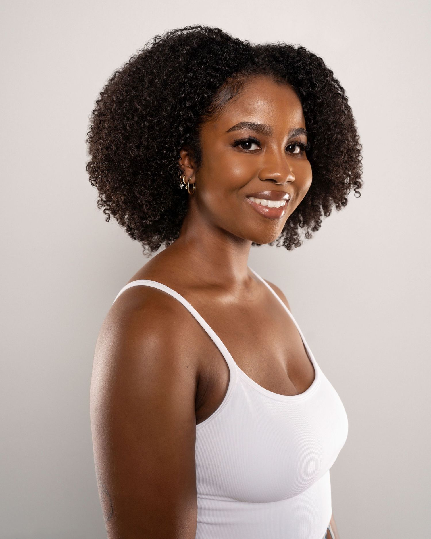 A young Black woman with natural curly hair smiles confidently, wearing a white camisole top against a neutral background.