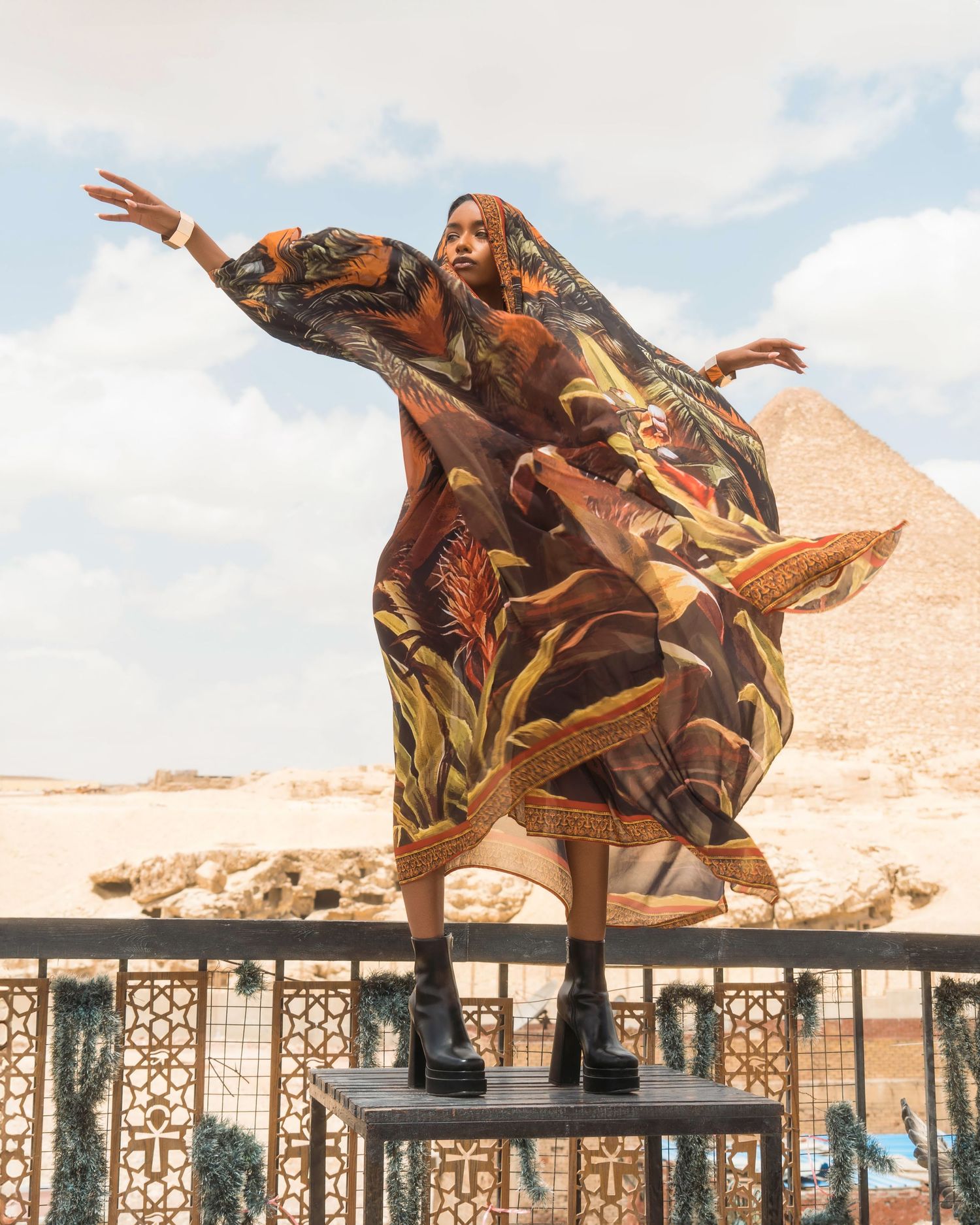 Person in floral dress performing dance moves on a balcony with desert landscape backdrop.