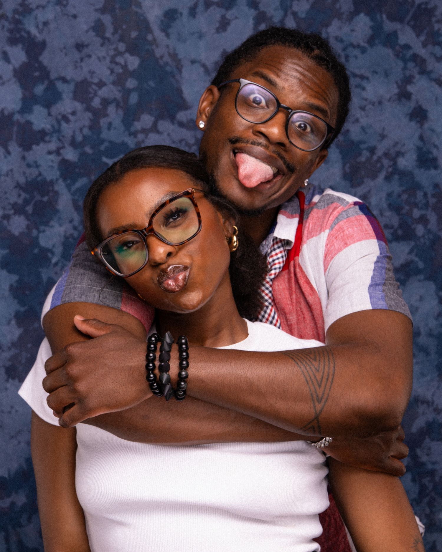 Two friends posing playfully together, making funny faces, wearing glasses, in front of a blue backdrop.