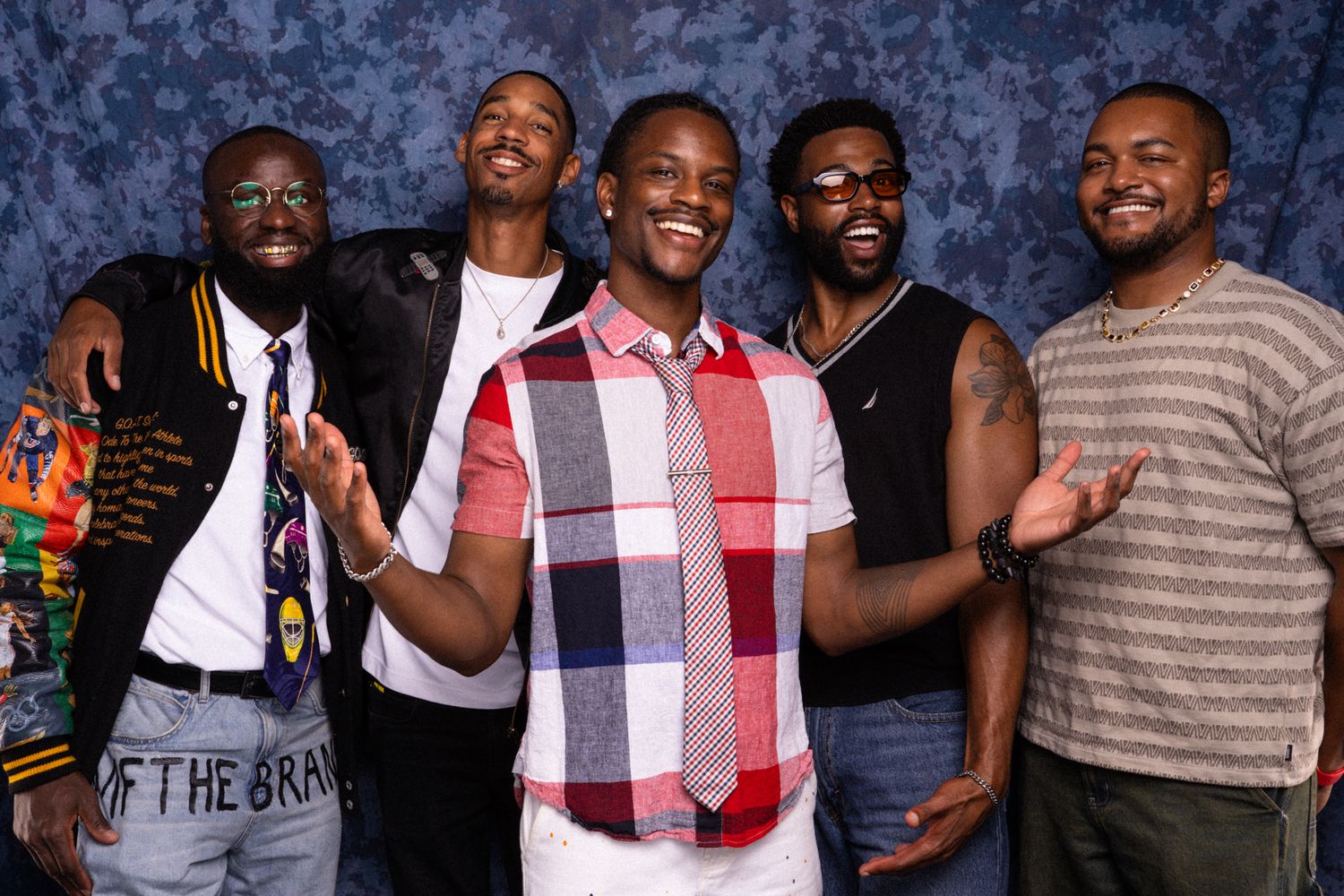 Five young men smiling and posing together in front of a blue backdrop at a photo opportunity event.