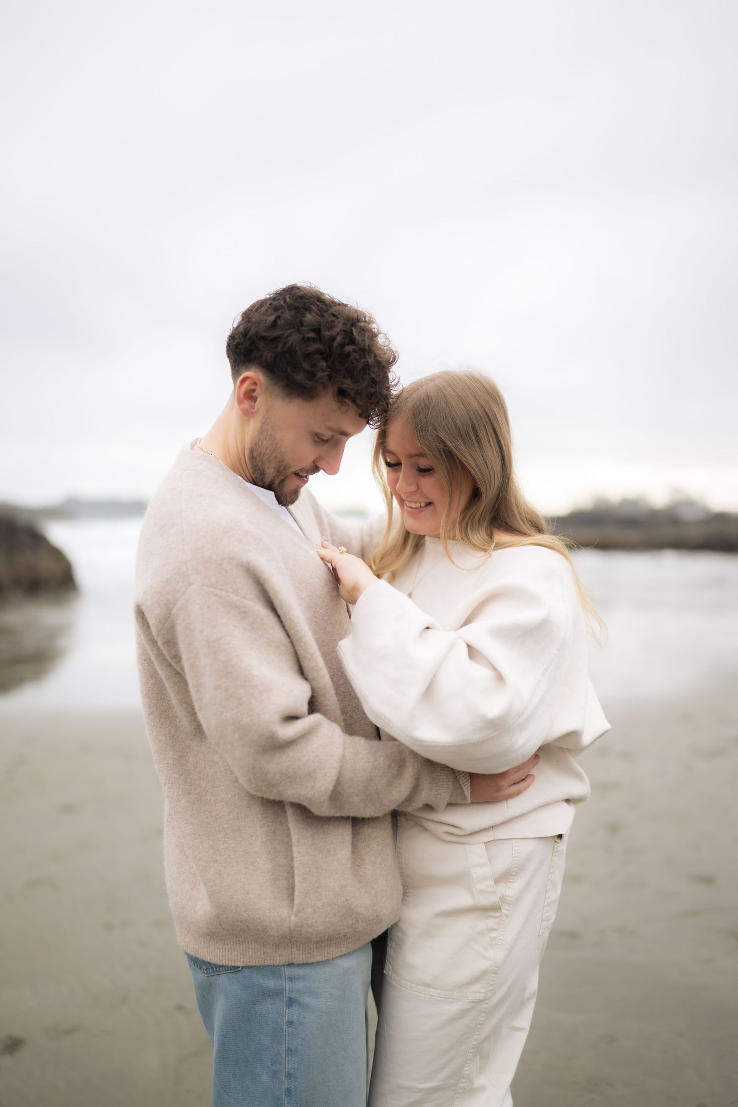 A romantic moment between a couple wearing cream colored sweaters on a cloudy beach.
