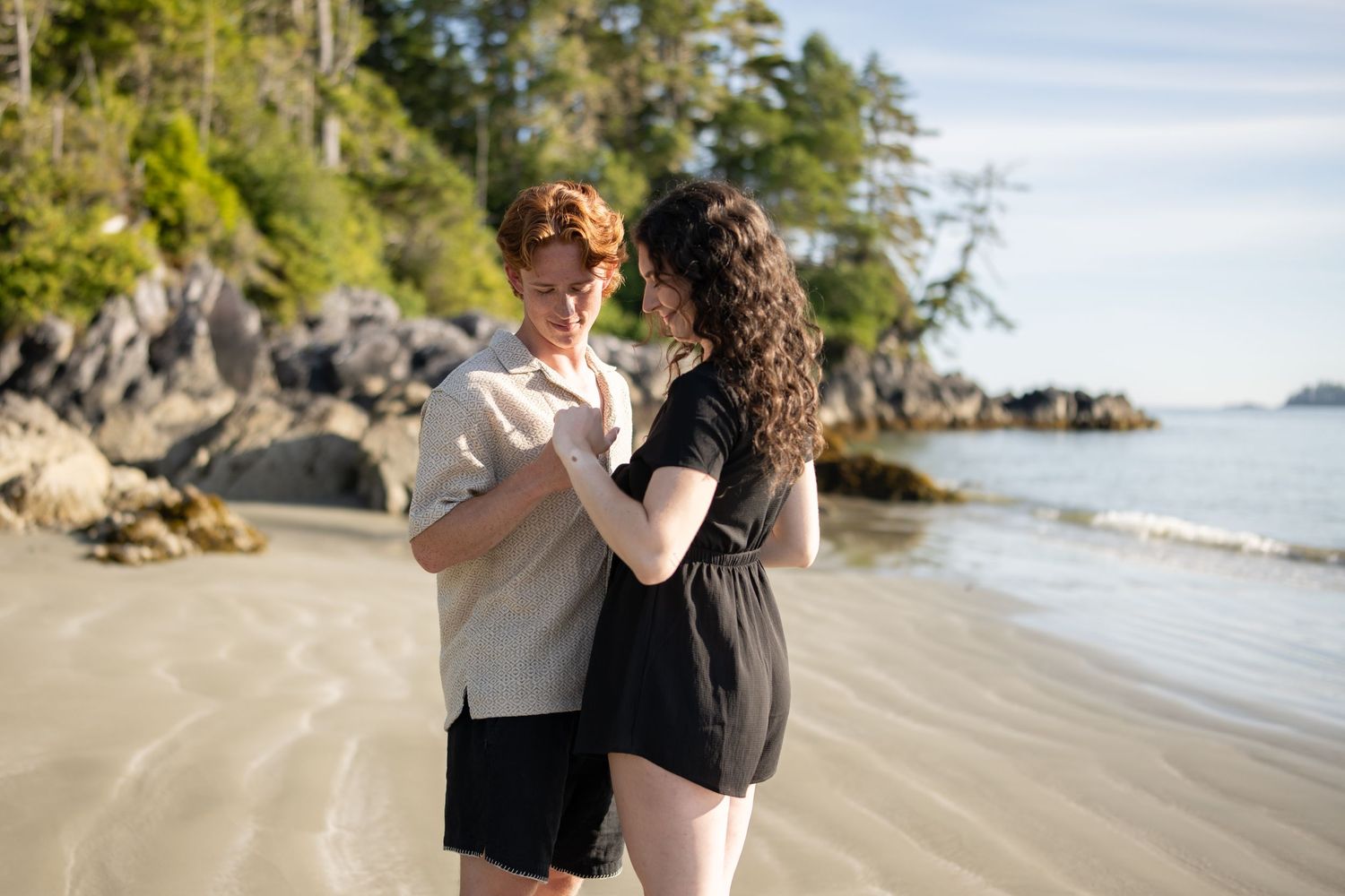 Couple embracing on a scenic beach with evergreen trees and rocky coastline.