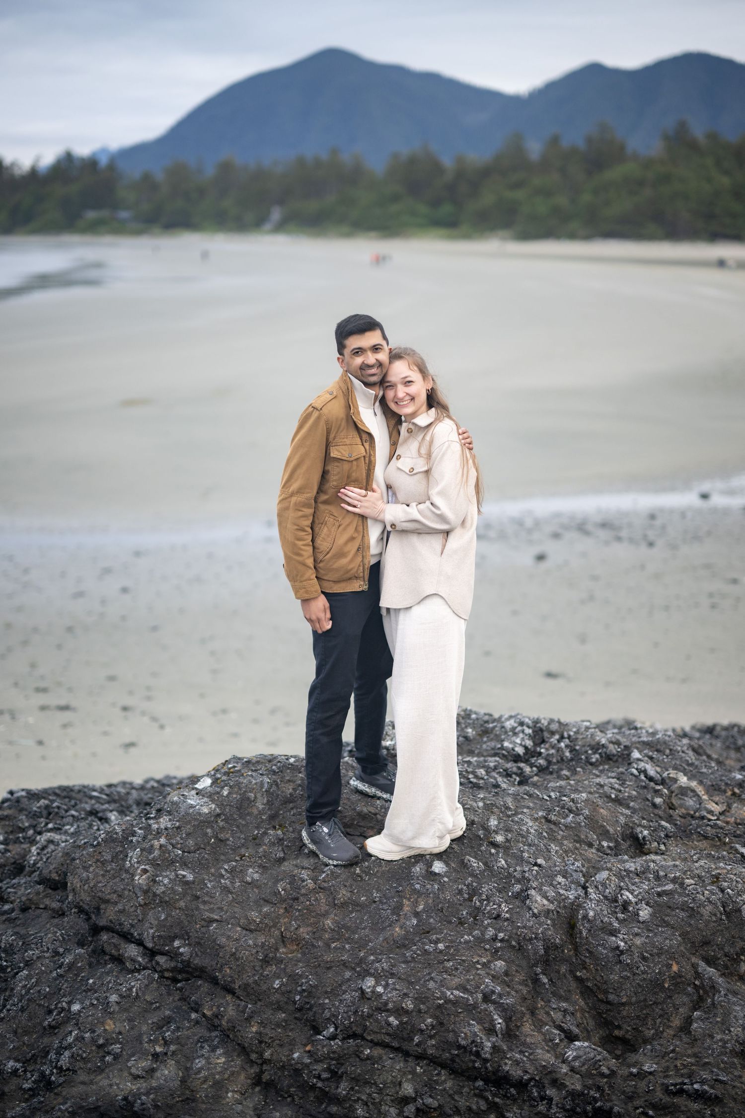 Couple standing on dark rocky outcrop with misty beach background.