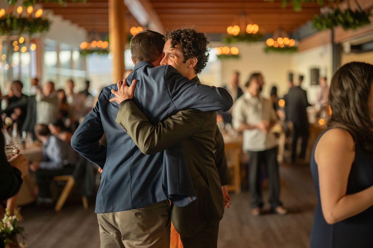 Wedding guests dance and celebrate on a wooden dance floor under warm lighting at an indoor reception venue.