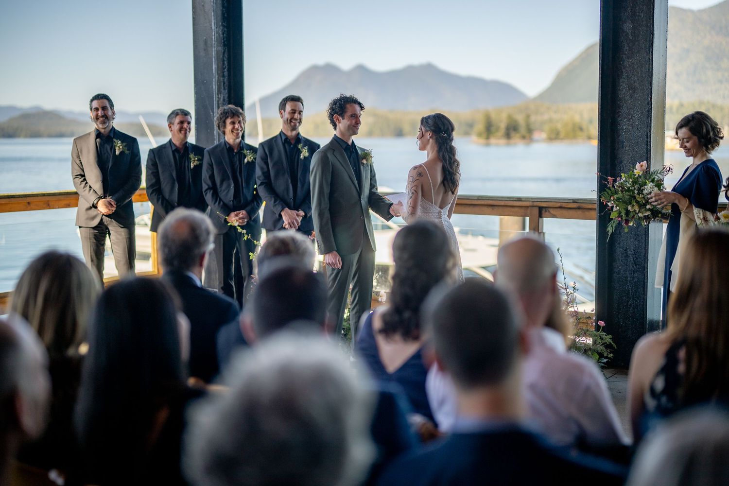 Wedding ceremony in progress at lodge with mountains and lake visible through windows.