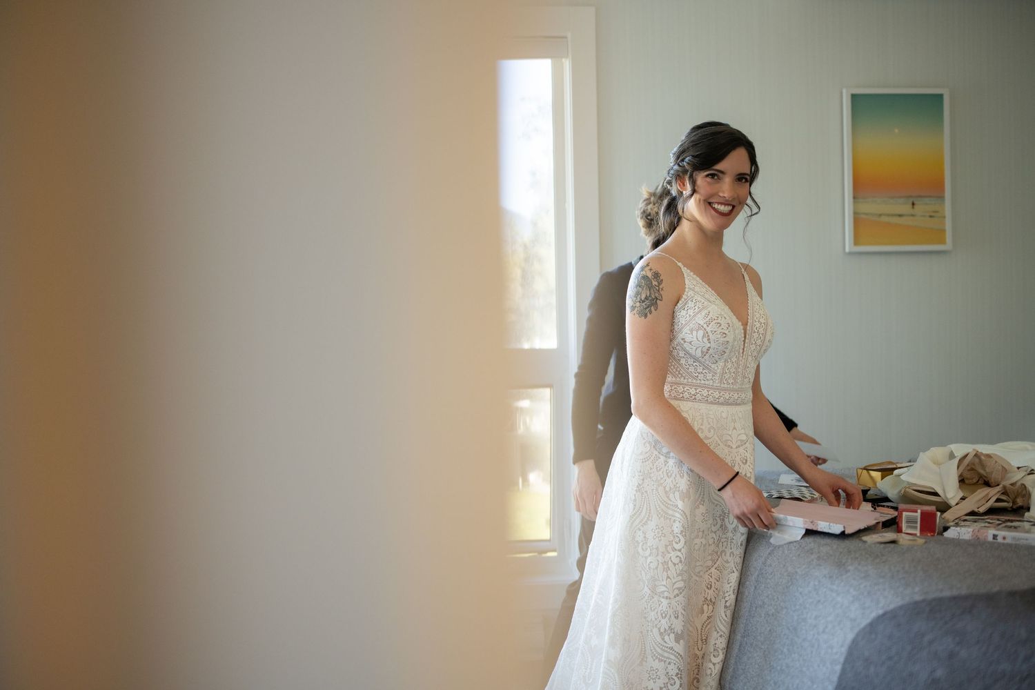 A white lace wedding dress being fitted in a sunlit room with a framed artwork on the wall.