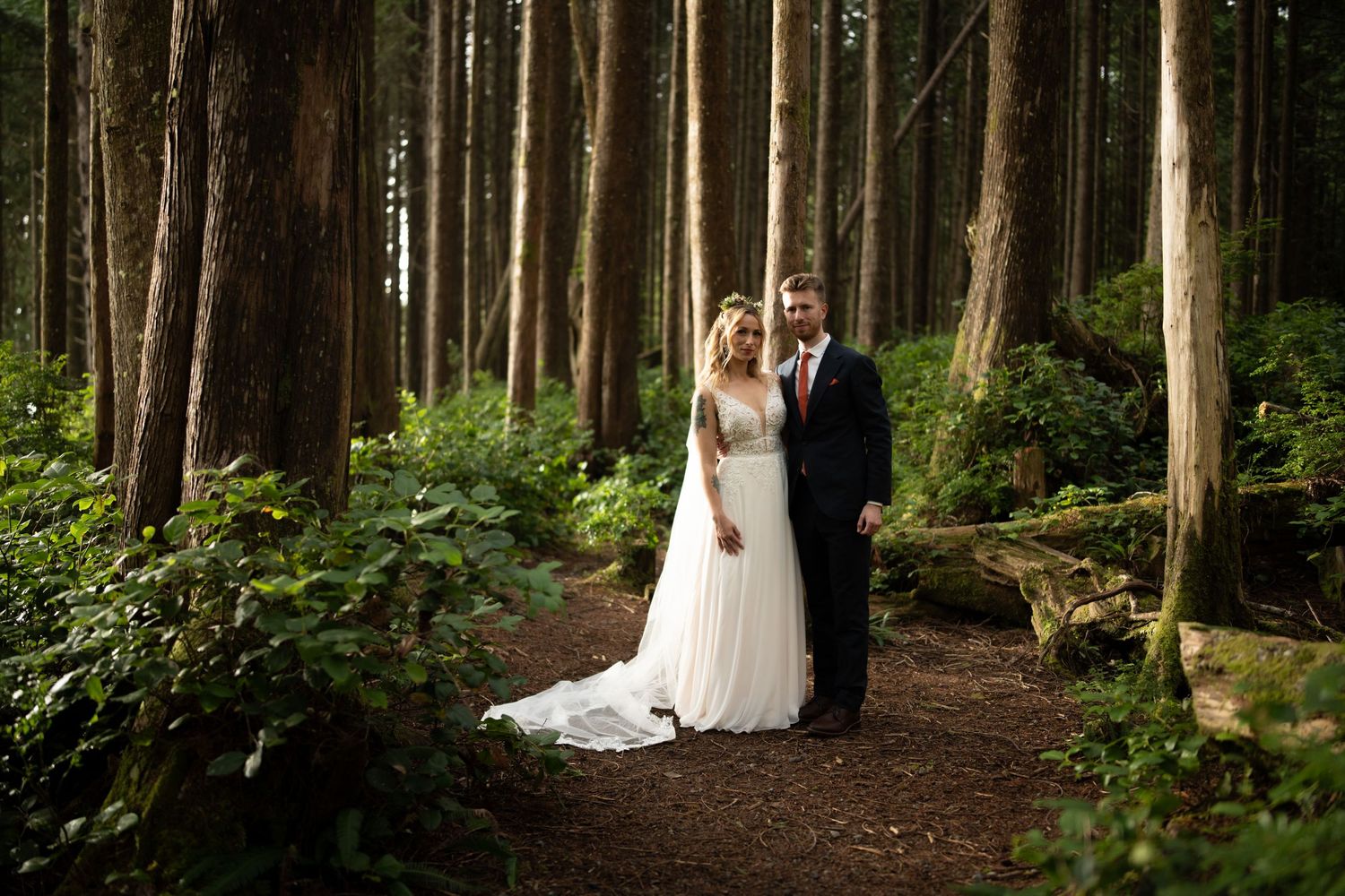 Wedding couple embraces on forest path surrounded by towering evergreens.