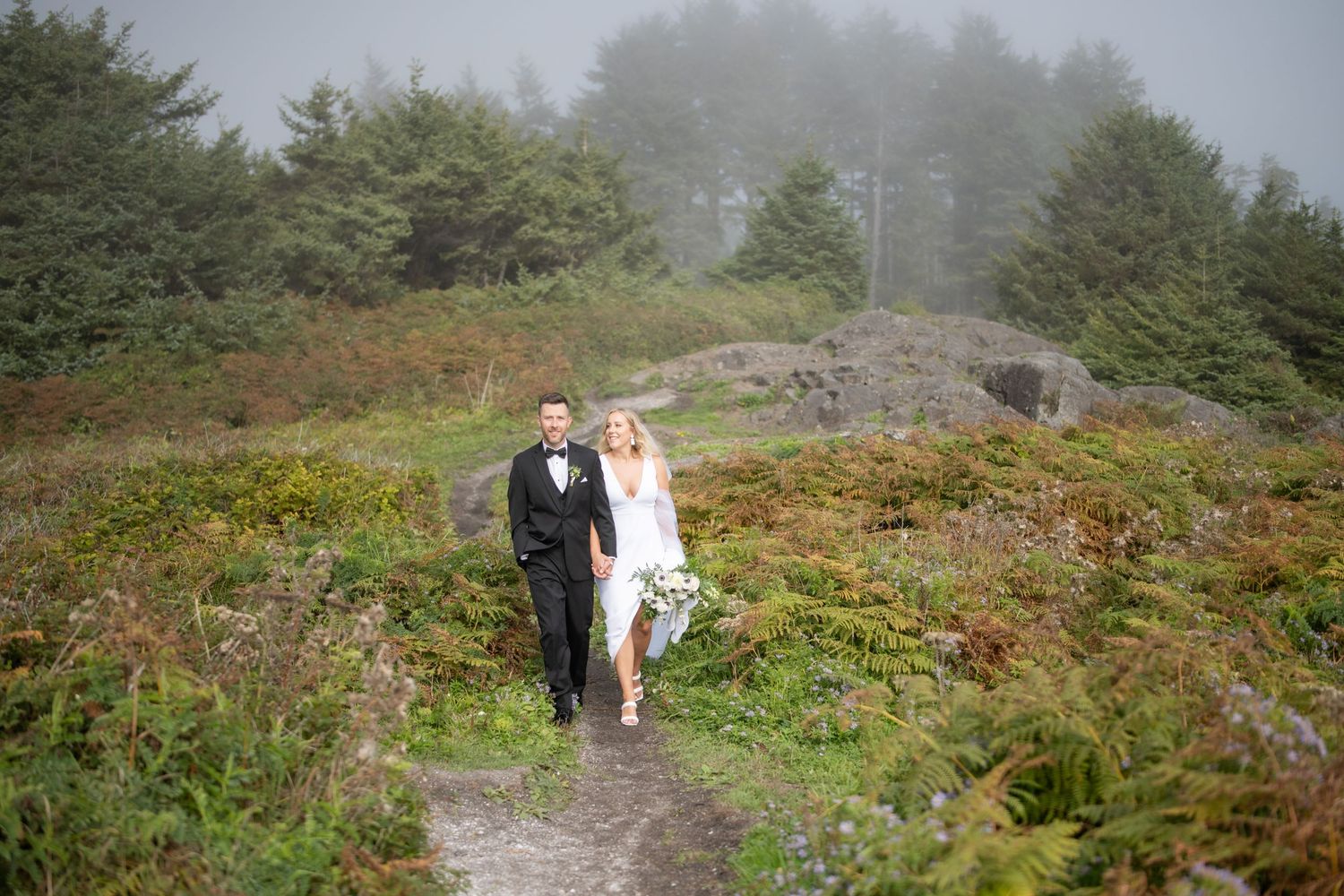 A couple in formal attire walking down a scenic mountain path on a foggy day.