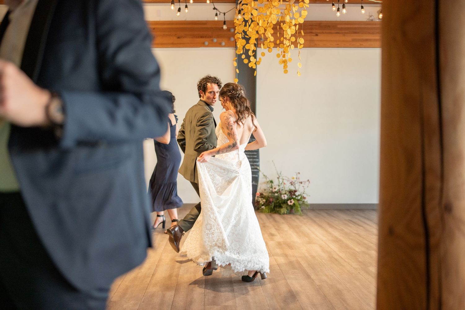 A festive wedding reception dance floor with guests celebrating under rustic wooden beams and twinkling lights.