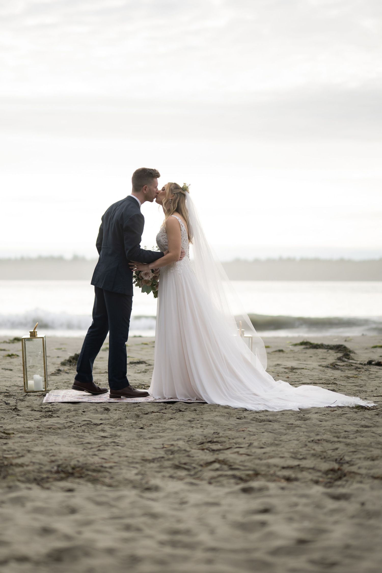Wedding couple shares intimate moment on sandy beach with overcast skies.