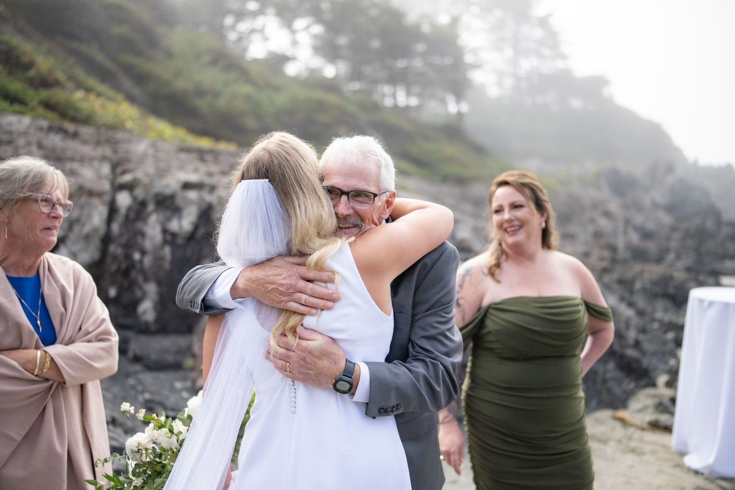 Heartfelt hugs shared during beach wedding ceremony with rocky coastline backdrop.