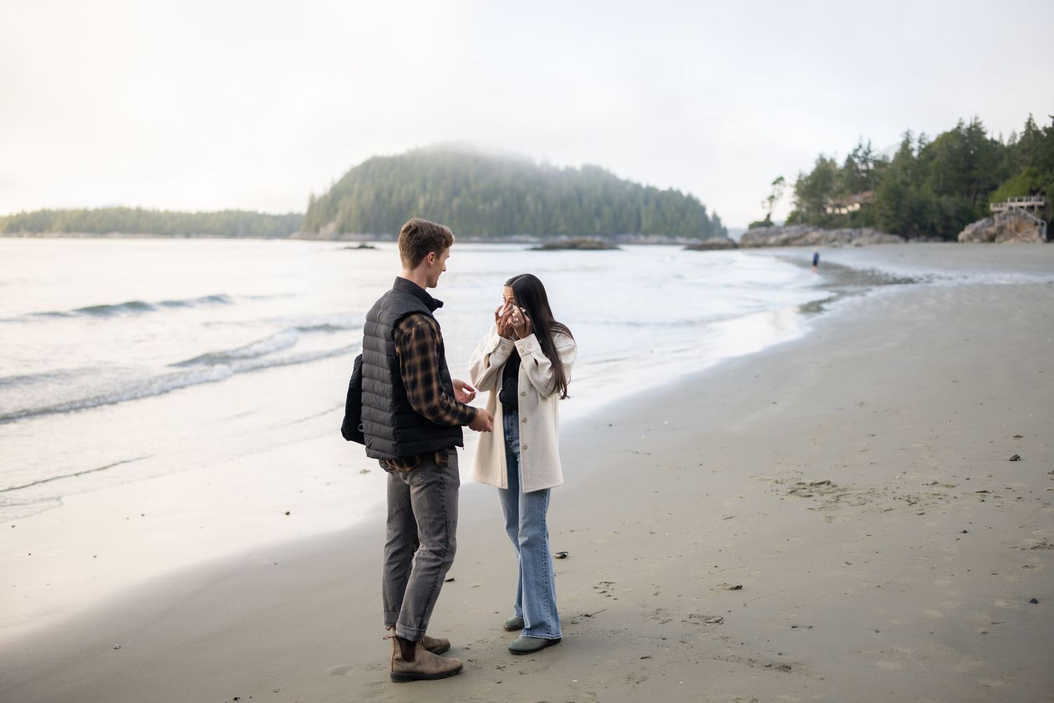 Foggy beach scene with evergreen trees and mountains visible in the background.
