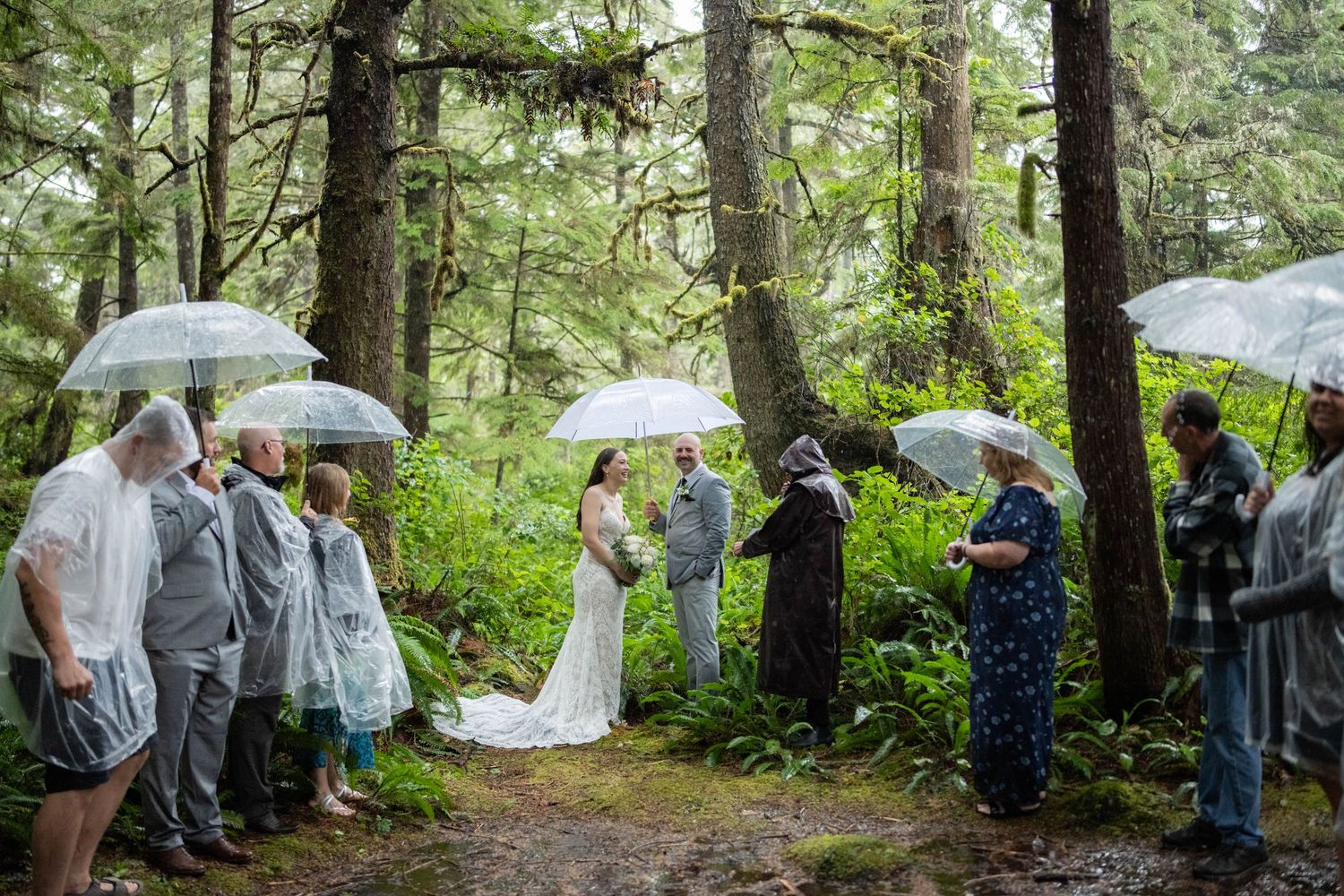 Wedding ceremony guests gather under umbrellas in a misty forest clearing.