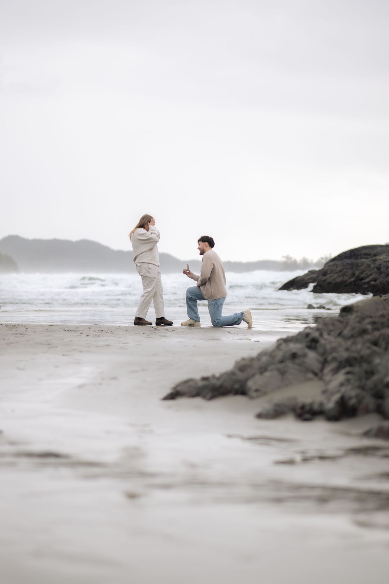 Dramatic beach proposal scene with rocky coastline and overcast sky in muted colors.