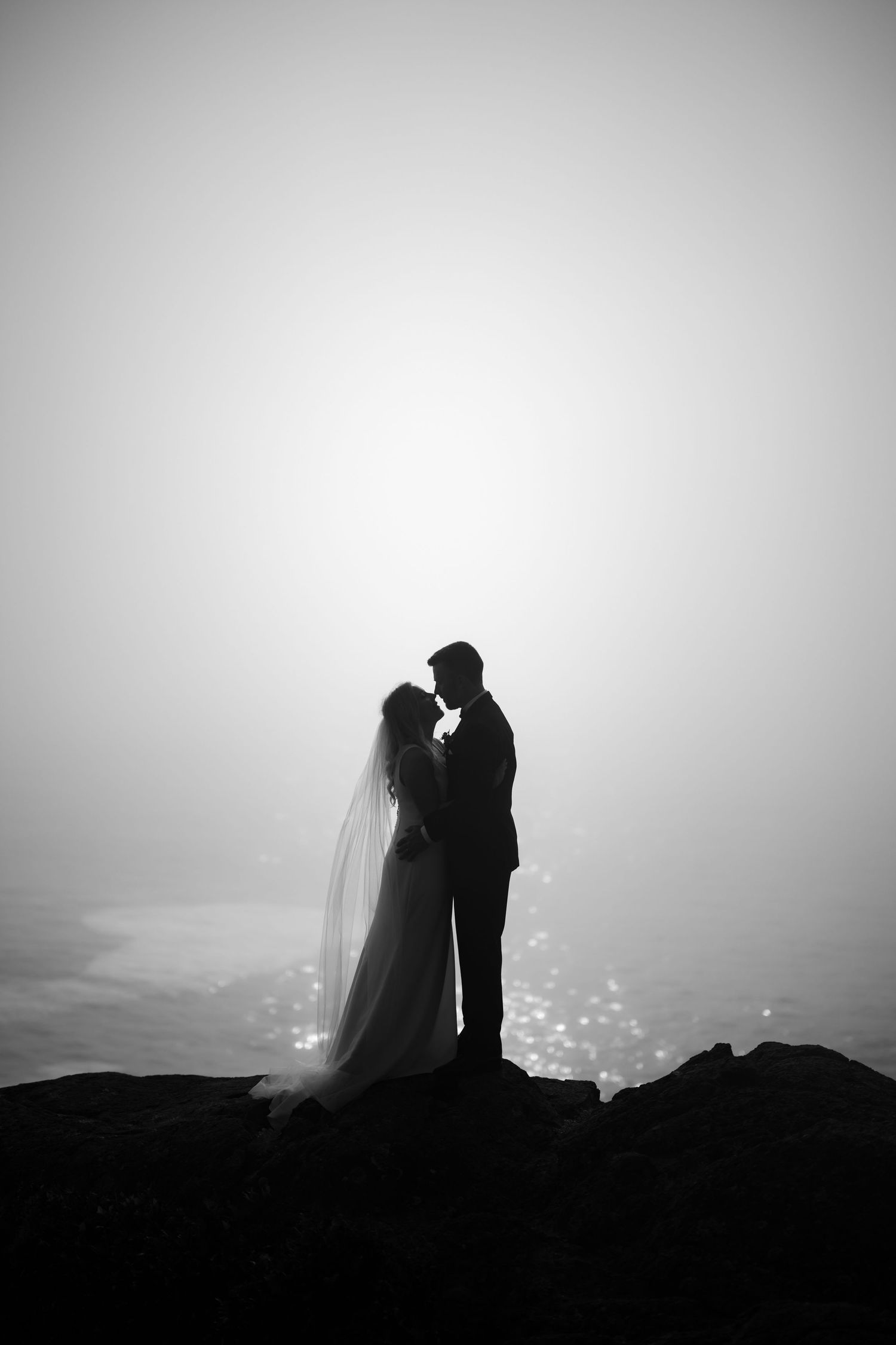 Silhouetted wedding couple embraces on clifftop against misty backdrop at dusk.