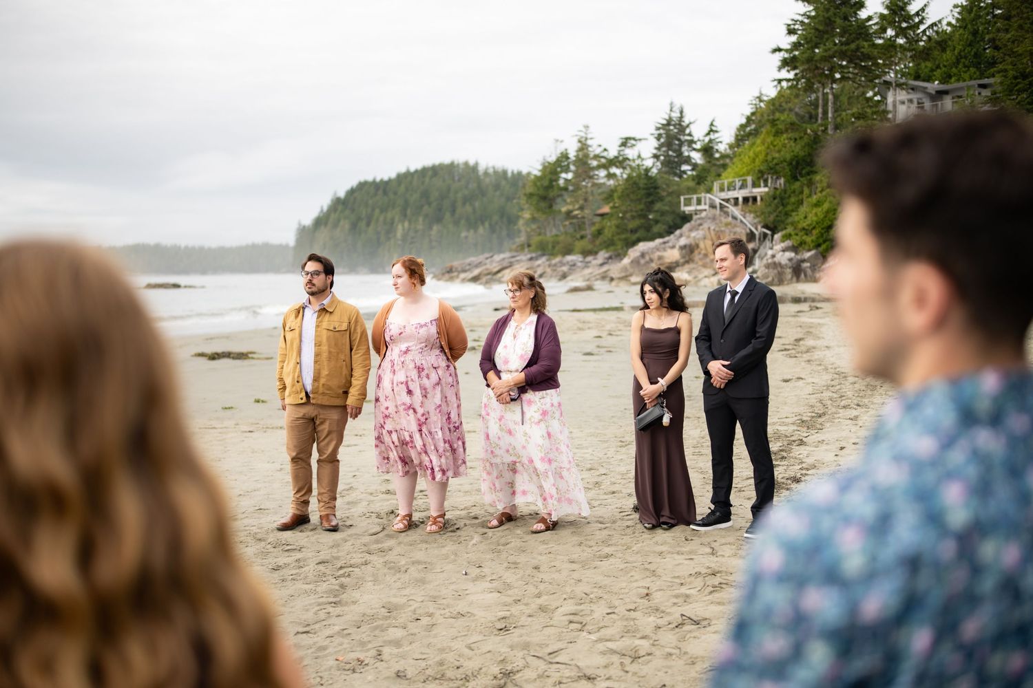 Wedding party stands on scenic beach with evergreen trees and mountains in background.