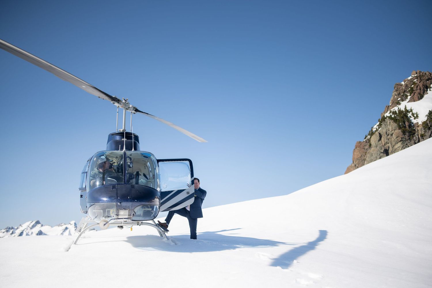 Helicopter landing on snowy mountain peak against bright blue sky.