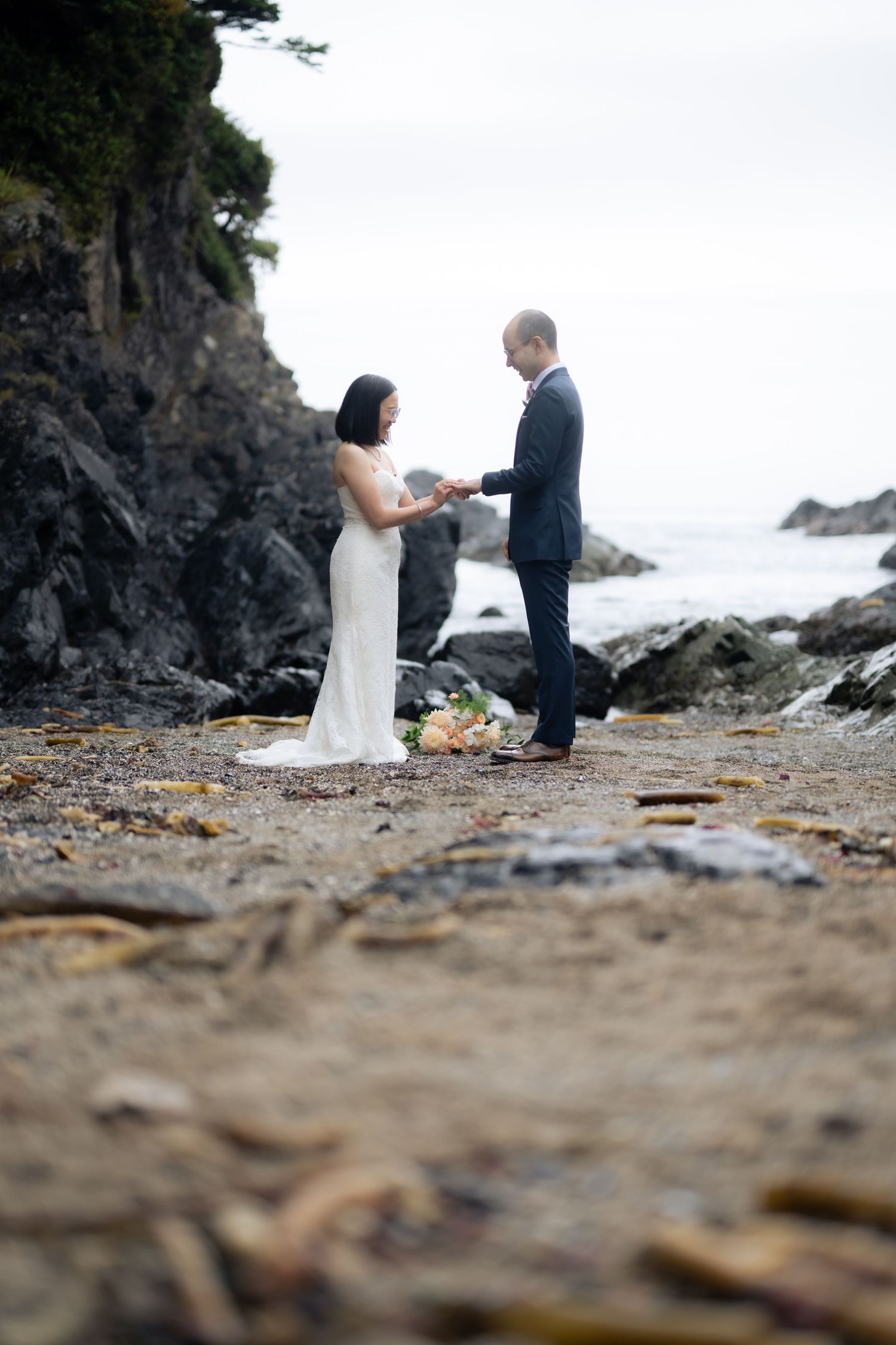 A couple exchanges vows during an intimate beach wedding ceremony with rocky cliffs in the background.