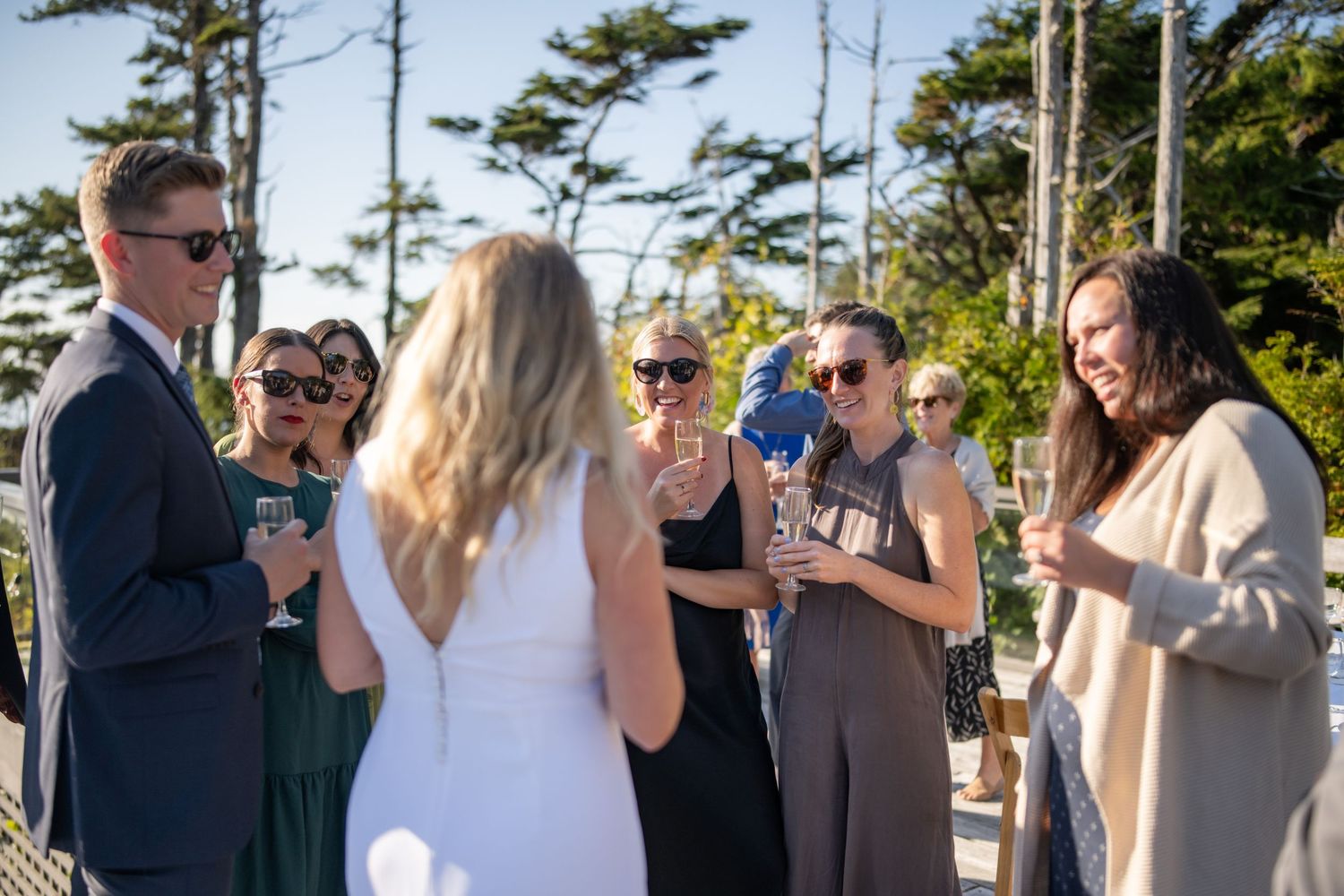 Wedding guests gather for cocktail hour on an outdoor deck surrounded by tall pine trees.