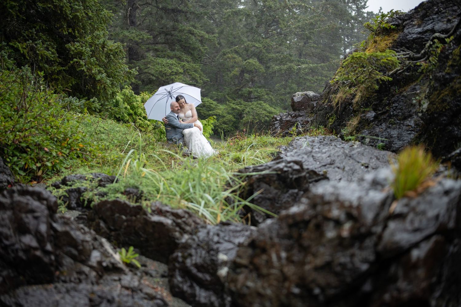 A couple shares an intimate moment on rocky terrain surrounded by lush greenery.