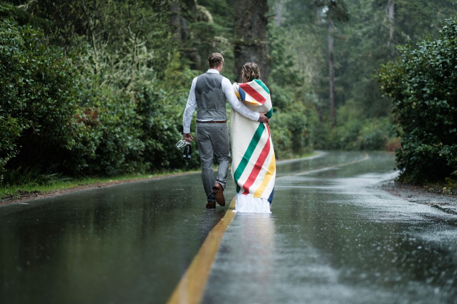 A couple walks down a wet road carrying a colorful striped blanket.