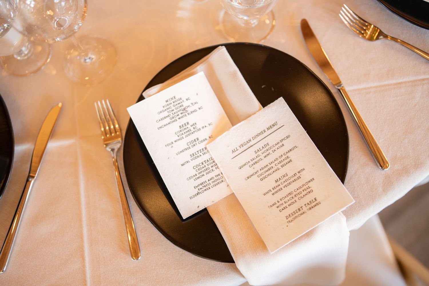 Wedding reception place settings with menus on black plates and gold flatware.