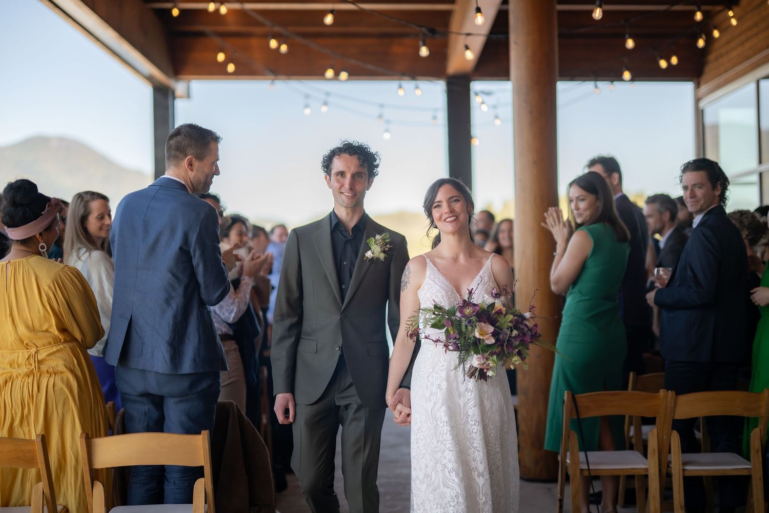 Wedding celebration in a rustic covered pavilion with wooden beams and string lights overhead.