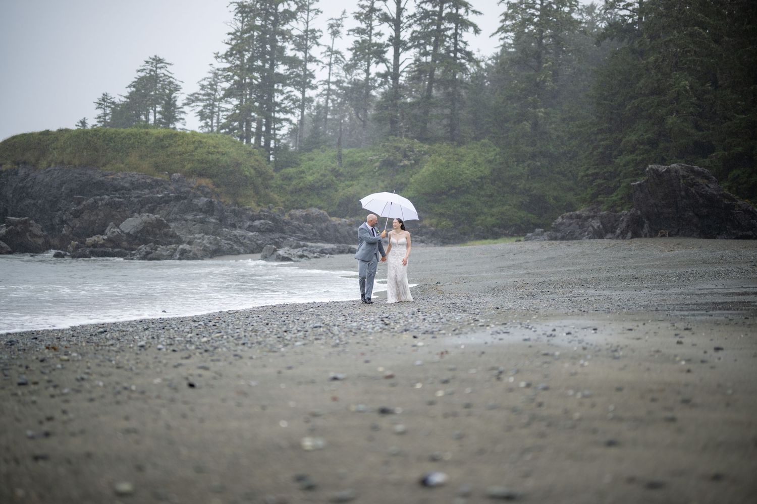 Overcast beach scene with evergreen trees and rocky shoreline in the distance.