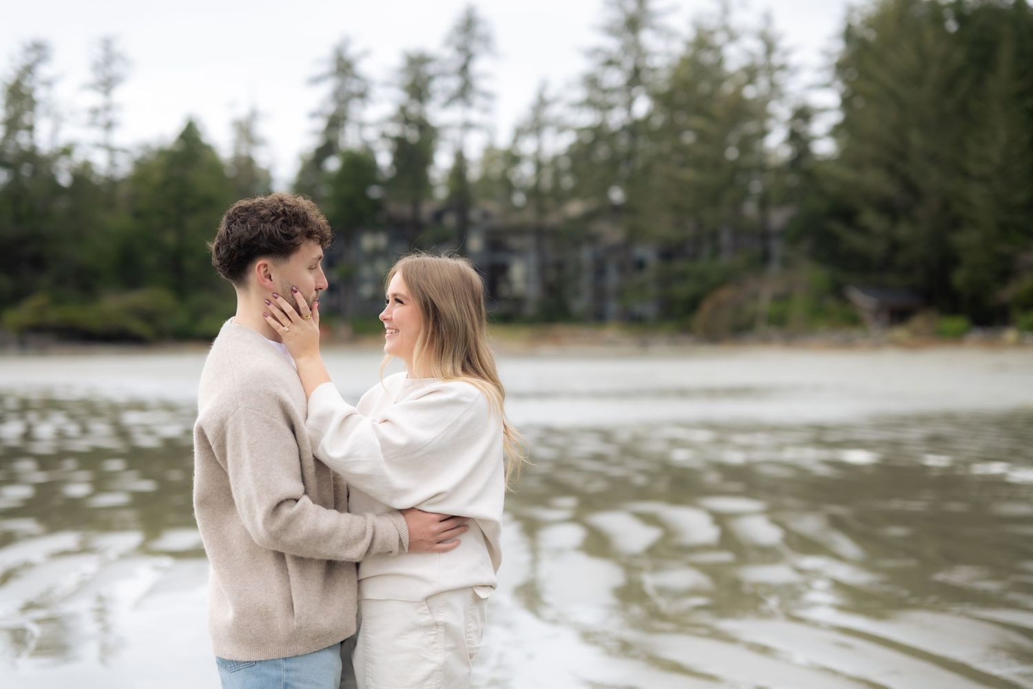 A tender moment between two people in neutral sweaters by the water's edge.